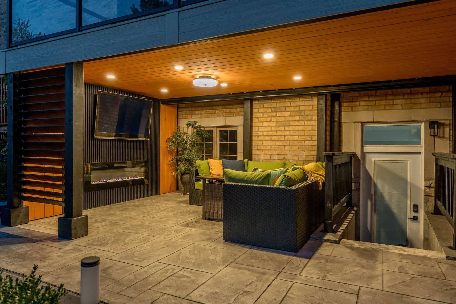 Covered hardscape patio beneath a composite deck with black-framed glass railings in Bradford