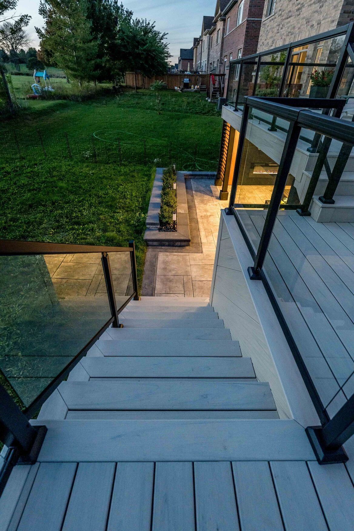View down wide composite deck stairs with black-framed glass railings toward a lit Bradford patio
